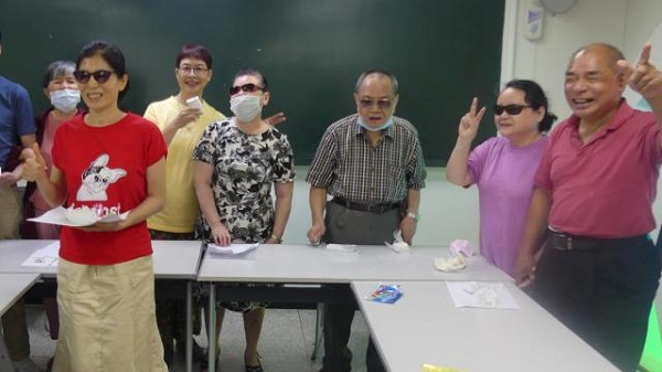 一群視障的爺爺奶奶們在教室裏圍著桌子向鏡頭比YA